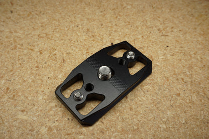 Dead Center Arca Plate in black anodized aluminum, angled top view showing the knurled face, center mounting screw, and two adjustment screws